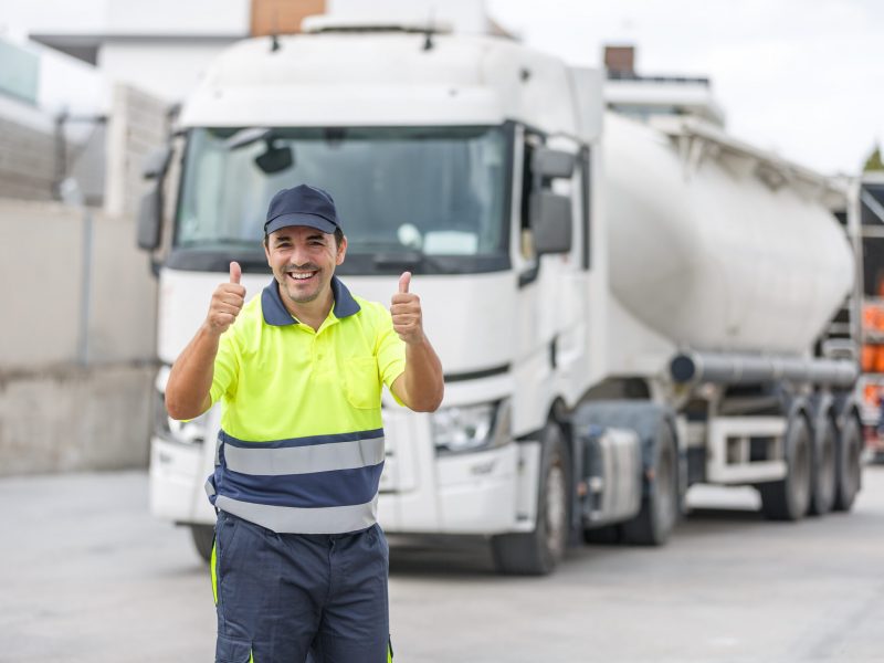 Cheerful male mechanical engineer in casual uniform and cap smiling and showing double thumb up gesture while standing and approving tanker truck